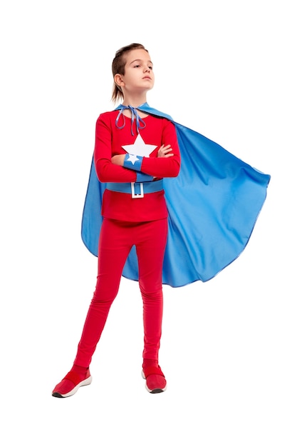 Ernstige jongen van volledige lengte in superheldenkostuum met wuivende cape die de armen kruist en wegkijkt terwijl hij tegen wit staat