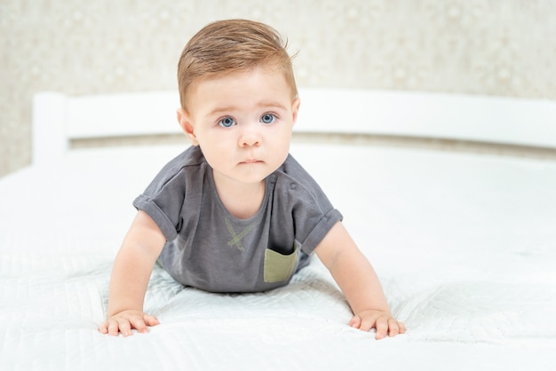 Ernstige kleine baby die op het bed kruipt op een wit laken in de slaapkamer van de ouders, baby die naar de camera kijkt