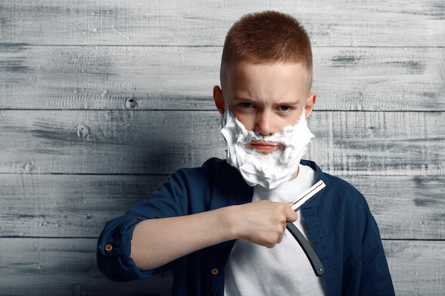 Ernstige kleine jongen met scheerschuim op zijn gezicht houdt een scheermesje in de studio.