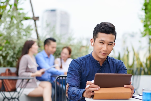 Ernstige knappe Aziatische zakenman zittend aan tafel op terras en kijken naar presentatie op digitale tablet