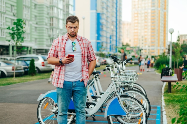 Ernstige man met een baard in vrijetijdskleding staat op de achtergrond van een rij deelfietsen en gebruikt een smartphone Man huurt een fiets via een mobiele applicatie