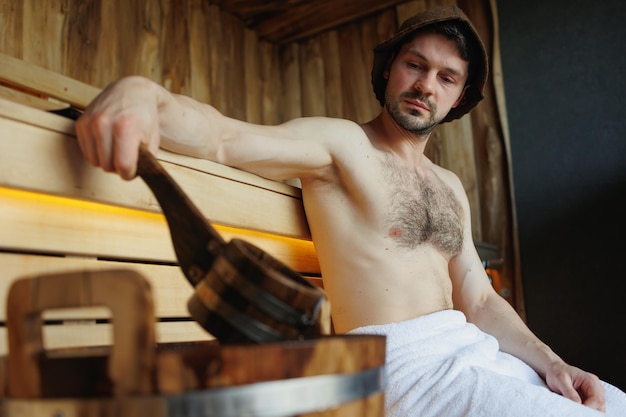 Ernstige man rust in een sauna en haalt water uit een emmer met een pollepel