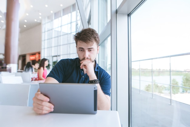 Ernstige man zit aan de tafel bij het raam en kijkt gericht op de tablet. Werk aan de tablet in het café