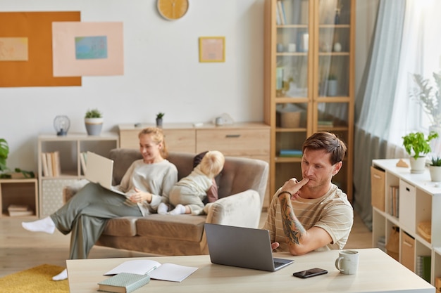 Ernstige man zit aan de tafel en concentreert zich op zijn online werk op laptop met zijn gezin thuis