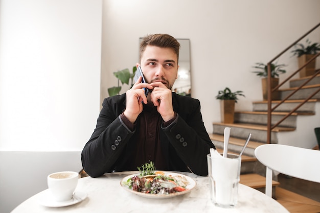 Ernstige man zit aan een tafel in de buurt van een bord met salade en een kopje koffie, praten aan de telefoon in het restaurant