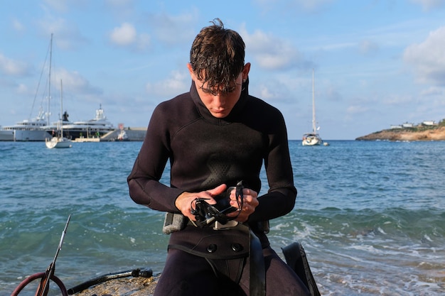 Ernstige mannelijke duiker in wetsuit zit op een rots bij een harpoen en kijkt naar beneden terwijl hij duikbril schoonmaakt voordat hij in de blauwe zee duikt