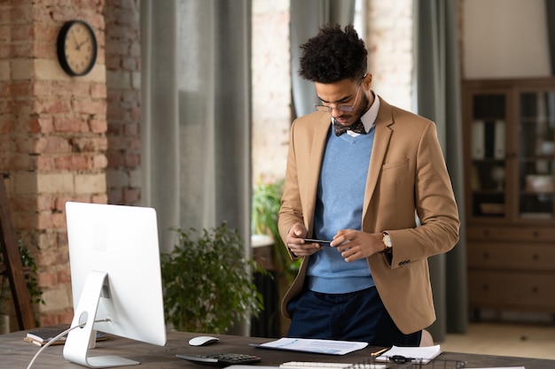 Ernstige modieuze jonge zakenman van gemengd ras met afro-kapsel die aan tafel staat met computer en papieren op telefoon fotografeert