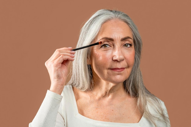 Ernstige mooie oude blanke vrouw met grijs haar past cosmetica toe met penseel geïsoleerd op bruine achtergrond