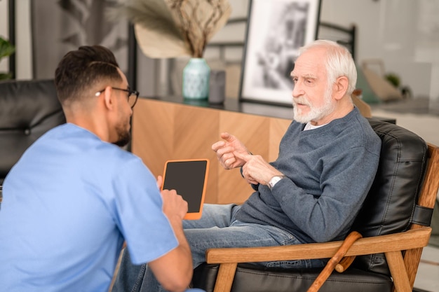 Ernstige oude man zittend in de fauteuil voor zijn verzorger dingen op zijn vingers te tellen tijdens het gesprek