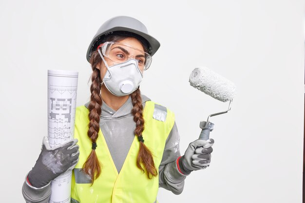 Ernstige professionele vrouwelijke architect die verantwoordelijk is voor het opstellen van gedetailleerde plannen voor structuur en het ontwikkelen van constructie draagt beschermend gezichtsmasker helmhandschoenen houdt blauwdruk verfroller vast