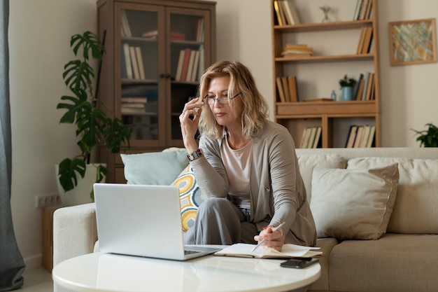 Ernstige rijpe blonde zakenvrouw in vrijetijdskleding zittend op de bank door kleine tafel voor laptop tijdens het netwerken in de woonkamer