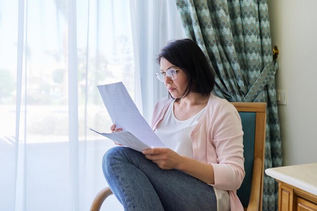 Ernstige rijpe vrouw die documenten leest die thuis in een leunstoel zitten