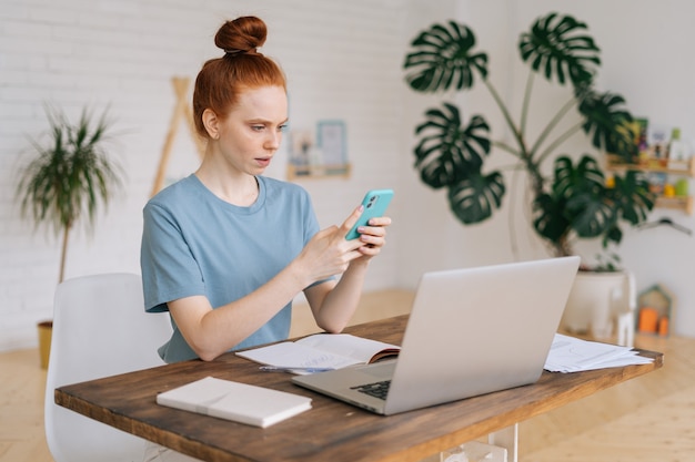 Ernstige roodharige jonge zakenvrouw gebruikt mobiele telefoon terwijl ze aan het bureau zit