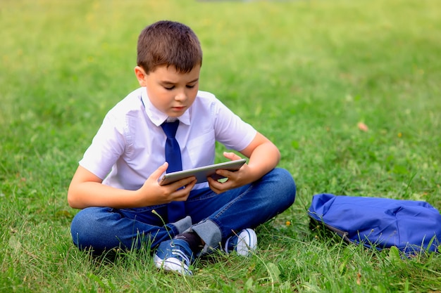 Ernstige schooljongen in een wit overhemd en een blauwe stropdas zit op groen gras tegen een blauwe lucht met witte wolken en kijkt naar een tablet.