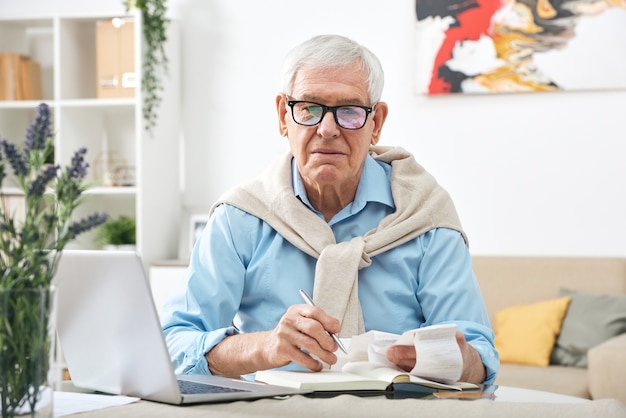 Ernstige senior man in bril kijken via betalingsrekeningen en notities maken in notitieblok zittend aan tafel