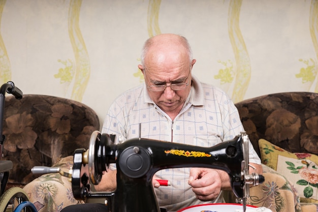 Ernstige Senior Man kijkt neer op de spoel van rode draad en rijgt de ouderwetse handmatige naaimachine thuis in de woonkamer in