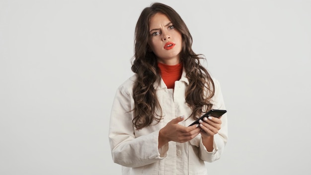 Ernstige stijlvolle brunette meisje geërgerd in de camera kijken met behulp van smartphone op witte achtergrond