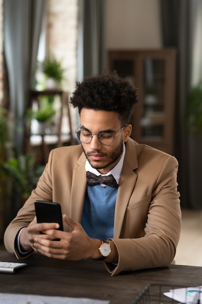 Ernstige stijlvolle jonge zakenman van gemengd ras met Afro-kapsel met een bril en een vlinderdas die aan het bureau zit en de telefoon controleert