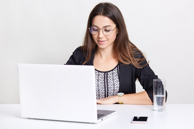 Ernstige tekstschrijver in zwarte kleding, gebruikt laptopcomputer voor werk en online communicatie, zit aan witte desktop, drinkt water, kijkt training webinar, doet aankoop.
