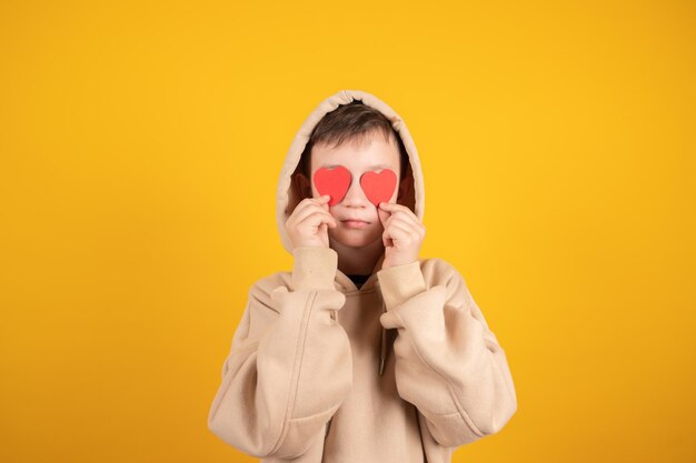 Ernstige tienerjongen die in handen twee kleine hartjes houdt Jonge jongen bedekt zijn ogen geïsoleerd op geel