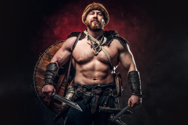 Ernstige Viking gekleed in lichte wapenrusting met een schild achter zijn rug houdt een zwaard en een bijl vast. Poseren op een donkere achtergrond met rood licht