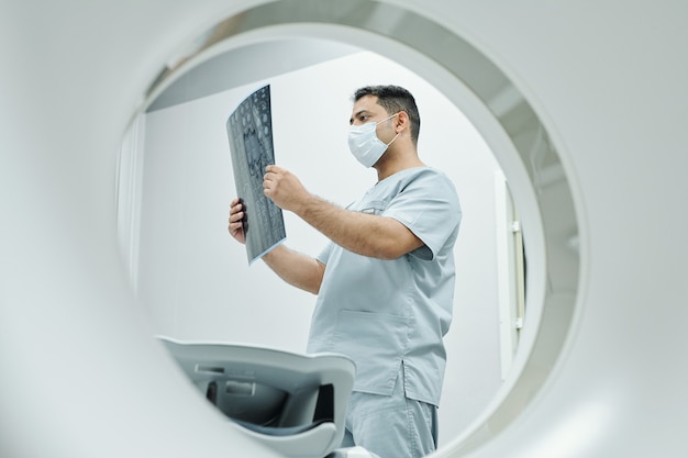 Ernstige volwassen radioloog van gemengd ras in masker en uniform kijkend naar röntgenfoto van het hoofd van de patiënt in een medische kantooromgeving in klinieken