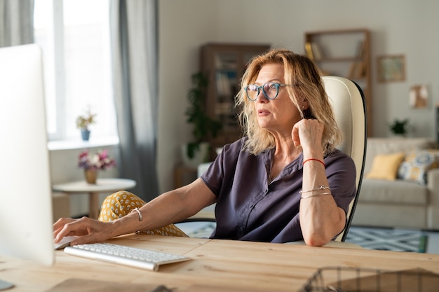 Ernstige volwassen vrouw of zakenvrouw in vrijetijdskleding zit door bureau achter computermonitor tijdens het werken op afstand thuis