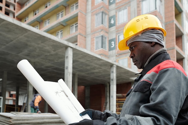 Ernstige volwassen zwarte man in werkkleding die naar de schets van de bouwstructuur kijkt