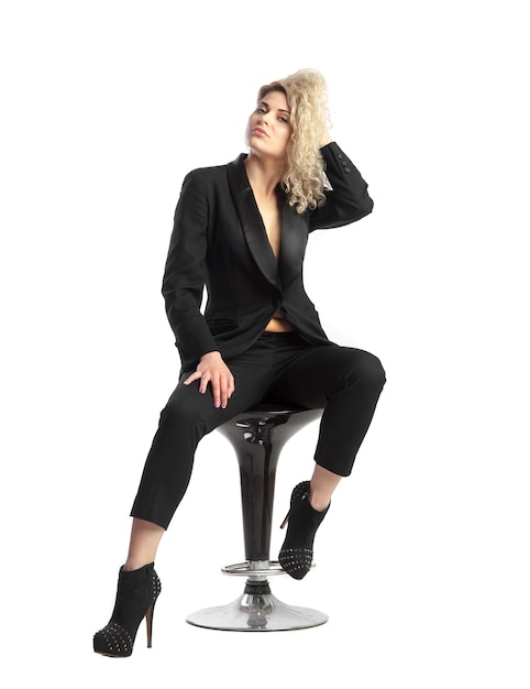 Ernstige vrouw met blond krullend haar zit op stoel in zwart pak op witte achtergrond