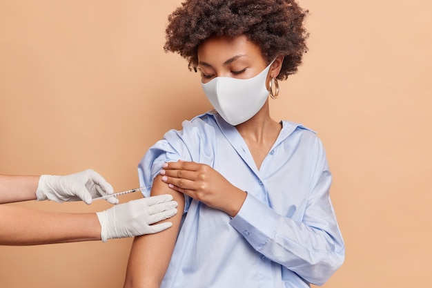 Ernstige vrouw met krullend haar draagt beschermend gezichtsmasker tegen virus draagt blauw shirt ontvangt inenting in schouder geïsoleerd op beige muur