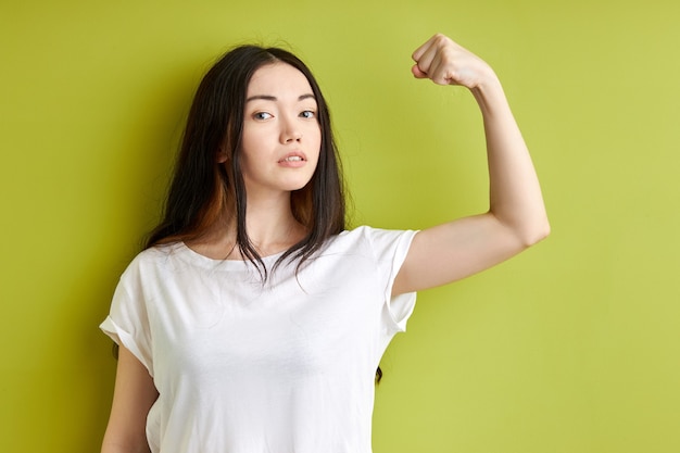 Ernstige vrouw met sterke spieren geïsoleerd op groene achtergrond, krachtige brunette vrouw in casual wit t-shirt kijkt naar de camera