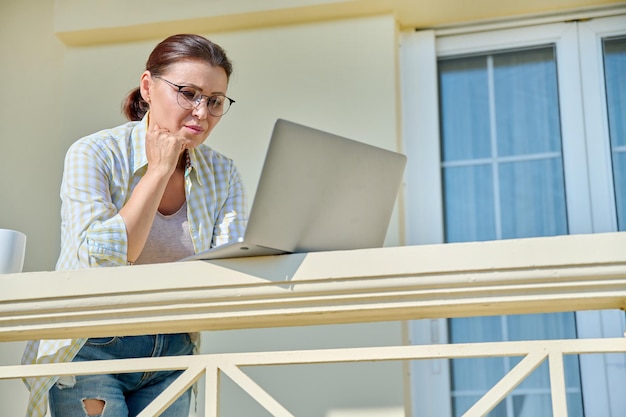 Ernstige vrouw van middelbare leeftijd met laptop op het balkon van het huis