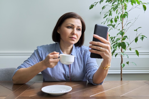 Ernstige vrouw van middelbare leeftijd met smartphone die thuis aan tafel zit