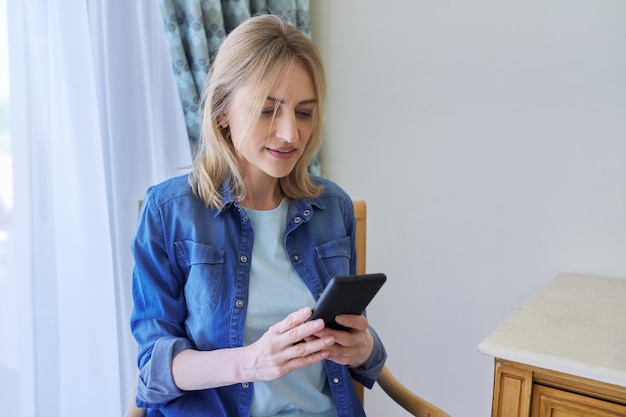 Ernstige vrouw van middelbare leeftijd met smartphone die thuis op een stoel zit