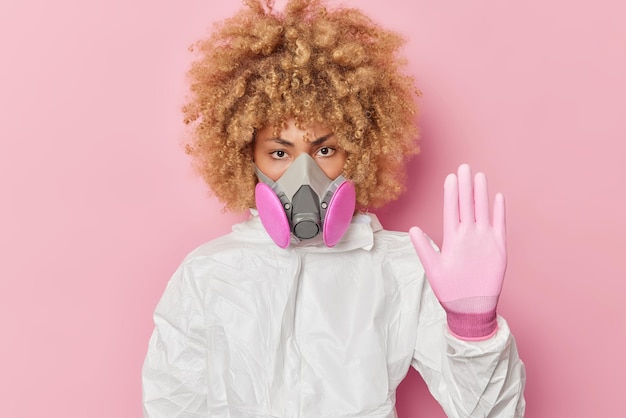 Foto ernstige vrouwelijke chemicus toont stopgebaar draagt beschermend pak gasmasker en handschoenen voorkomt biologisch gevaar geïsoleerd op roze achtergrond vrouwelijk model beschermt zichzelf tegen pandemie van het coronavirus