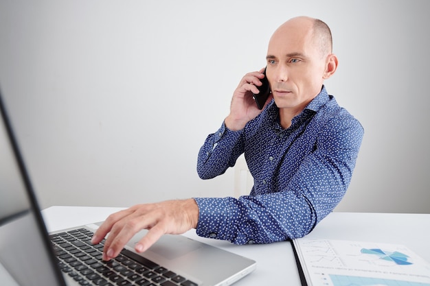 Ernstige zakenman van middelbare leeftijd die e-mails leest op het laptopscherm tijdens een telefoongesprek met zakenpartner of collega