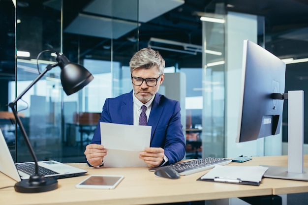 Ervaren grijsharige zakenman die op een computerpapierwerk op kantoor werkt en een brief van de bank leest