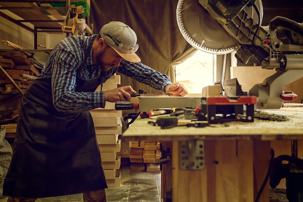 Ervaren timmerman in werkkleding en eigenaar van een klein bedrijf die in een houtbewerkingswerkplaats werkt en een cirkelzaag gebruikt om door een houten plank op tafel te zagen, is een hamer en veel gereedschap