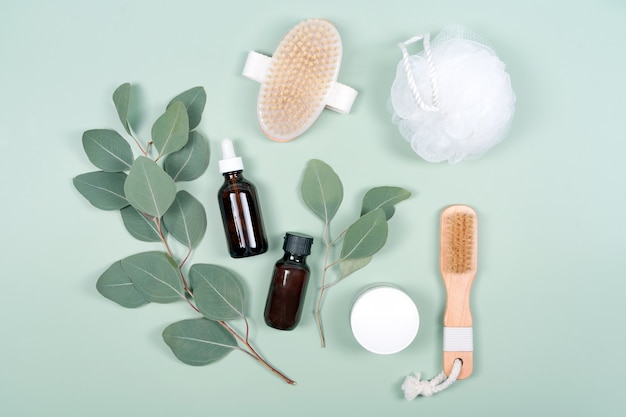 Essential oils, facial creme container and massage brushes with natural eucalyptus leaves on green background