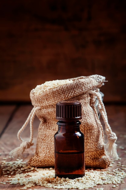 Essential sesame oil in a bottle of brown glass and sesame seeds in a bag of canvas on the old wooden background in rustic style toned image selective focus