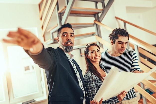 Estate agent is showing house to couple buyers, telling clients about home advantages. Interior designer is discussing renovation ideas with homeowners.