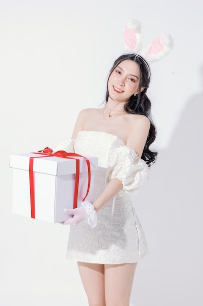 에스테르 축제 개념 귀여운 아시아 여성은 사랑스러운 얼굴을 가지고 있으며 완벽하고 깨끗한 피부와 날씬한 몸매를 가지고 있으며 외진 흰색 배경에 선물 상자를 들고 있는 보송보송한 토끼 귀를 가지고 있습니다.