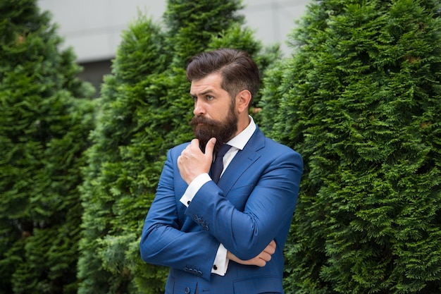 Esthete detail bearded man in jacket near cypress tree handsome man smartly dressed ceo walk in street Business man walking outdoor Businessman In Nature energy efficiency concept