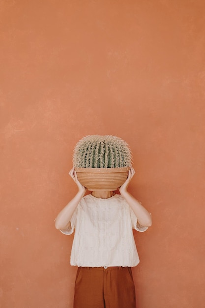 Esthetisch minimalistisch modeconcept Jonge mooie vrouw in gemberbroek en wit T-stuk houdt cactus tegen gembermuur