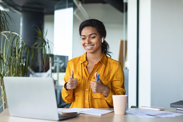 Eteaching Happy black female business tutor gesturing thumbs up to laptop computer webcamera sitting in office