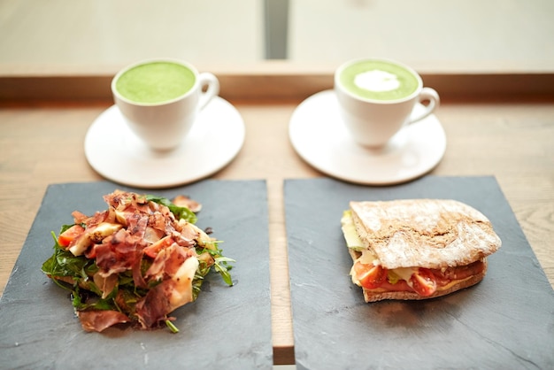eten, diner, haute cuisine en eetconcept - prosciutto ham salade met zalm panini sandwich op stenen borden en kopjes matcha groene thee latte in restaurant