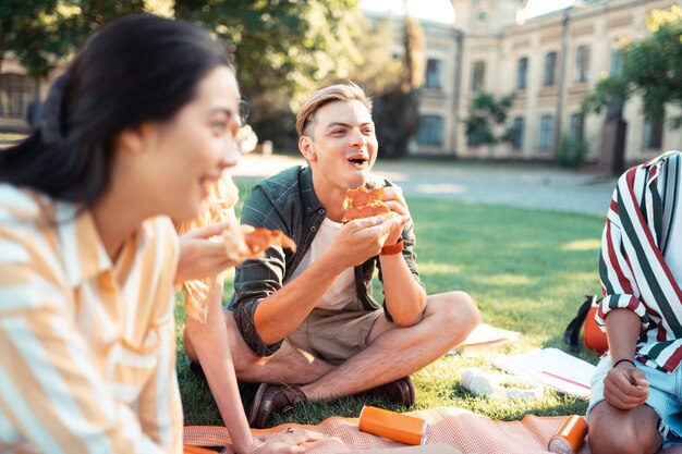 Eten met vrienden. Vrolijke jongen zit met zijn vrienden op het gras op de universiteitswerf lachend en zijn pizza etend.