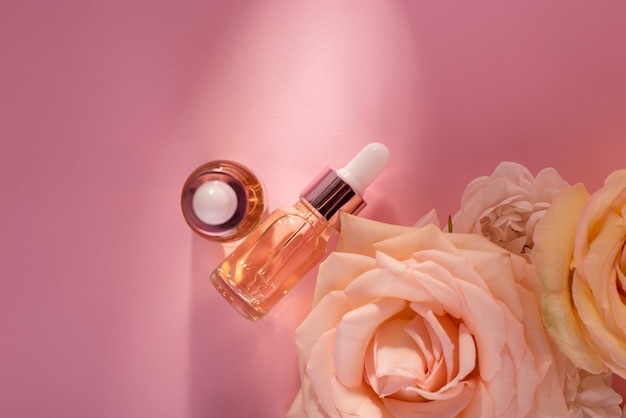 Etherische rozenolie in cosmetische fles in de buurt van verse roze bloemen tegen roze achtergrond. Bovenaanzicht cosmetica plat leggen