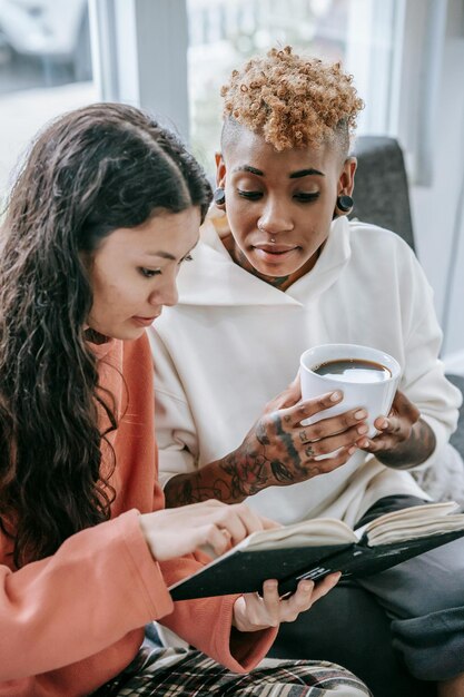 Photo ethnic female sharing textbook with black girlfriend sitting with cup of hot chocolate on couch at