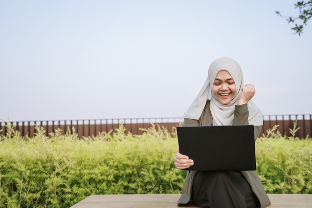 Euphoric winnaar Aziatische moslimvrouw in groene pak en werken op een computer in het park.